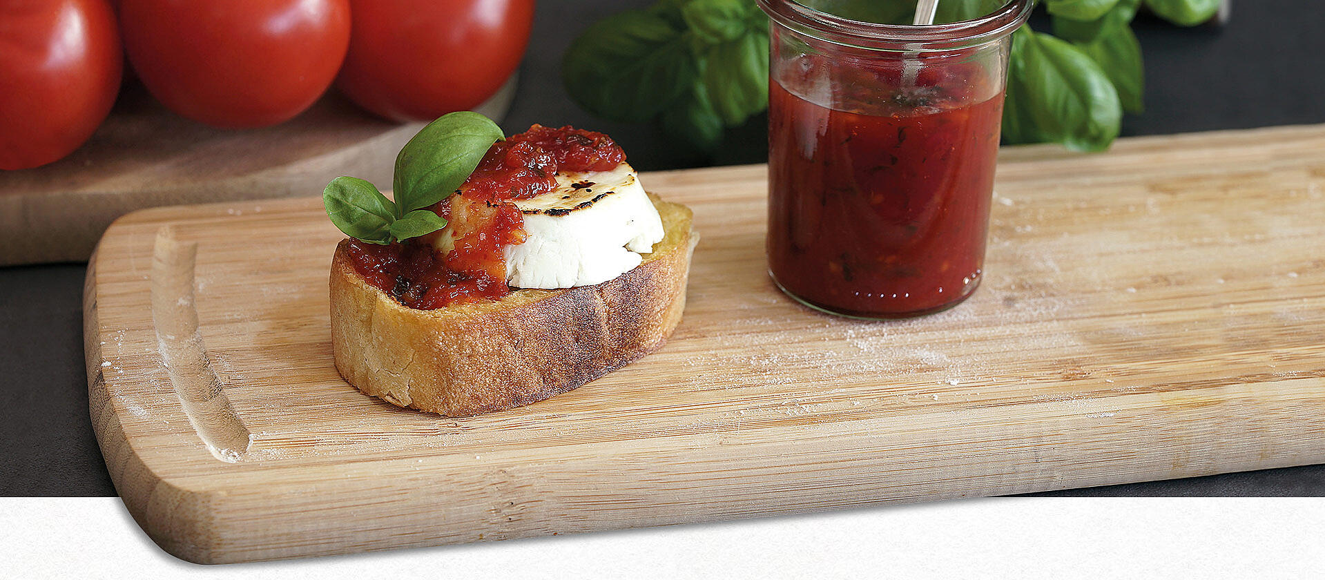 Auf einem Holzbrett liegt eine Scheibe Ciabatta mit gratiniertem Ziegenkäse und Tomatenmarmelade