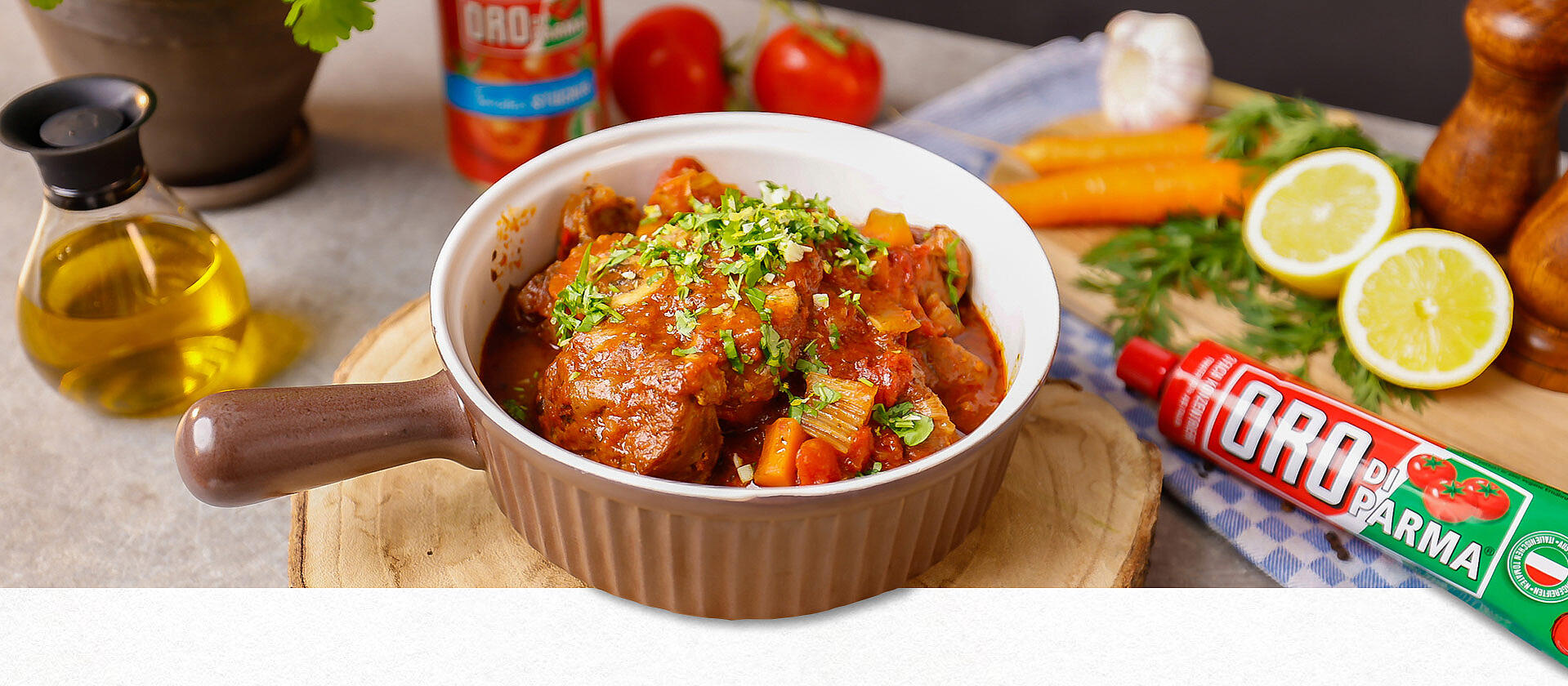 In einem Schmortopf ist Ossobuco alla milanese anrichtet. Der Topf steht auf einem runden Holzbrett. Im Hintergrund verschiedene frische Zutaten. 