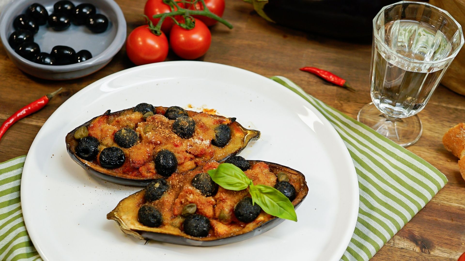 Auf einem weißen Teller liegen zwei Auberginenhälften. Beide sind gefüllt und überbacken. Im Hintergrund stehen ORO di Parma Tomaten, Oliven und frische Tomaten.