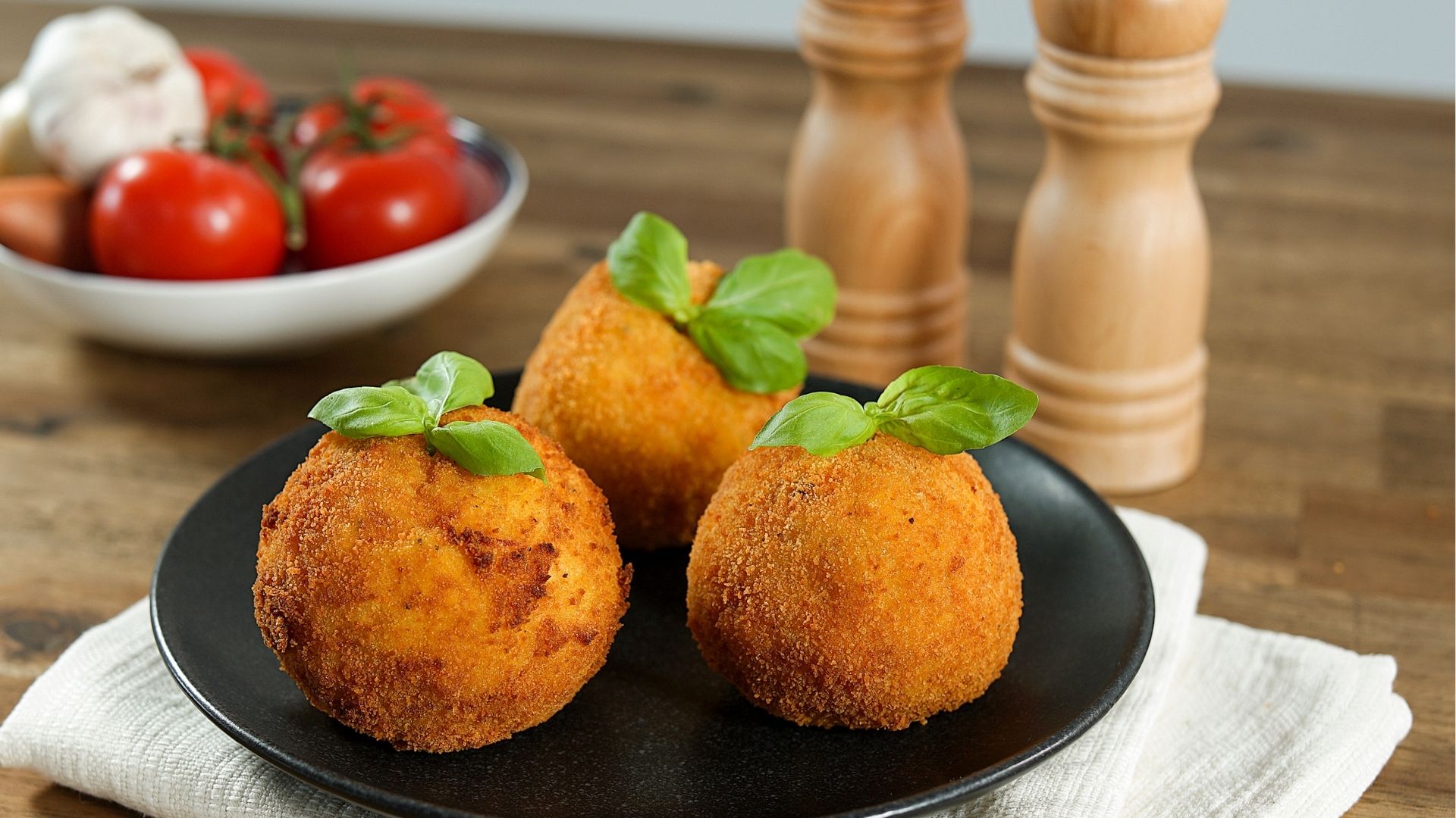 Arancini di riso - Reisbällchen auf einem schwarzen Teller. Im Hintergrund Salz- und Pfefferstreuer sowie frische Tomaten.