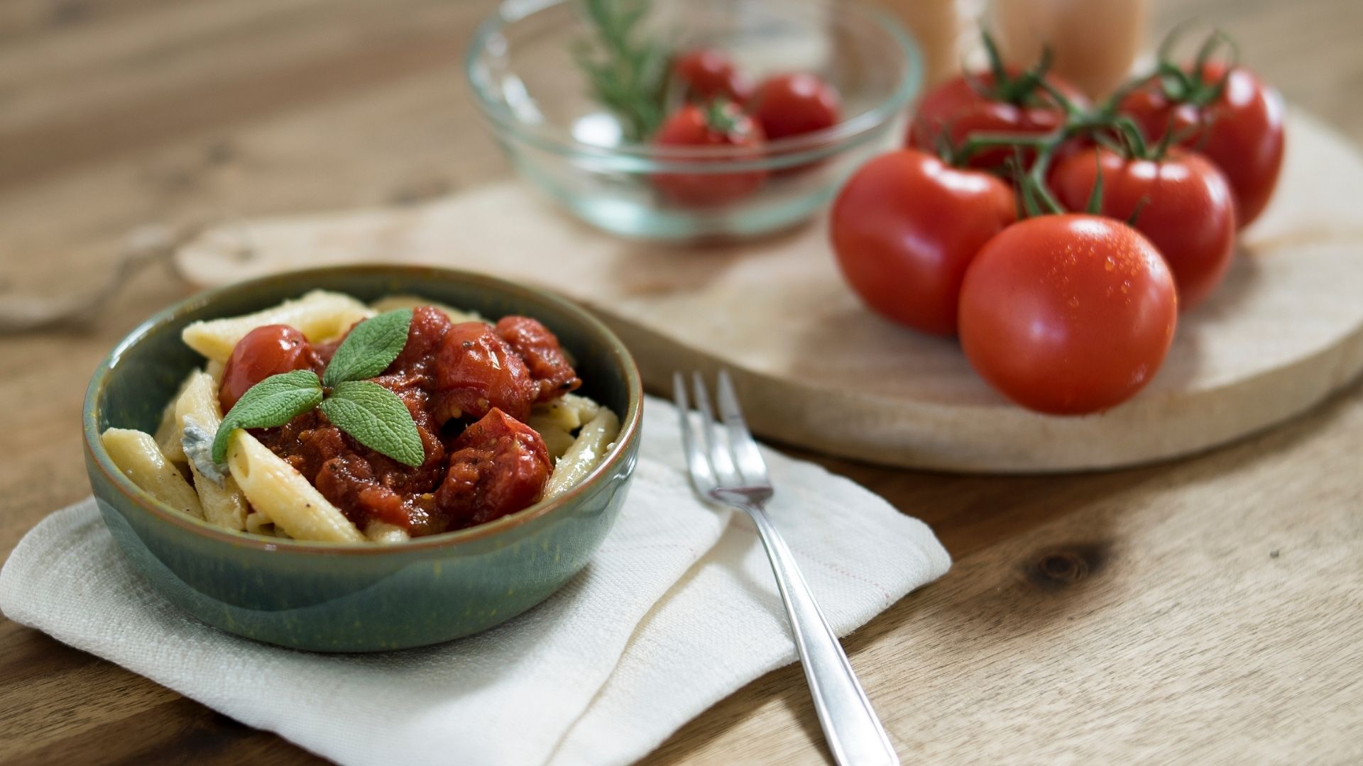 Eine Schüssel mit Penne Gorgonzola, links daneben ein Holzbrett mit Tomaten
