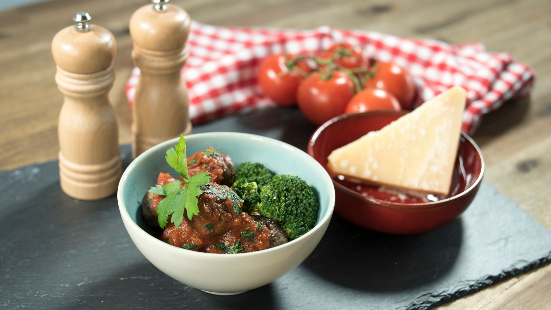Eine Schüssel mit Salami-Fleischbällchen in Tomatensugo mit Brokkoli, daneben eine Schüssel mit einem Stück Parmesan