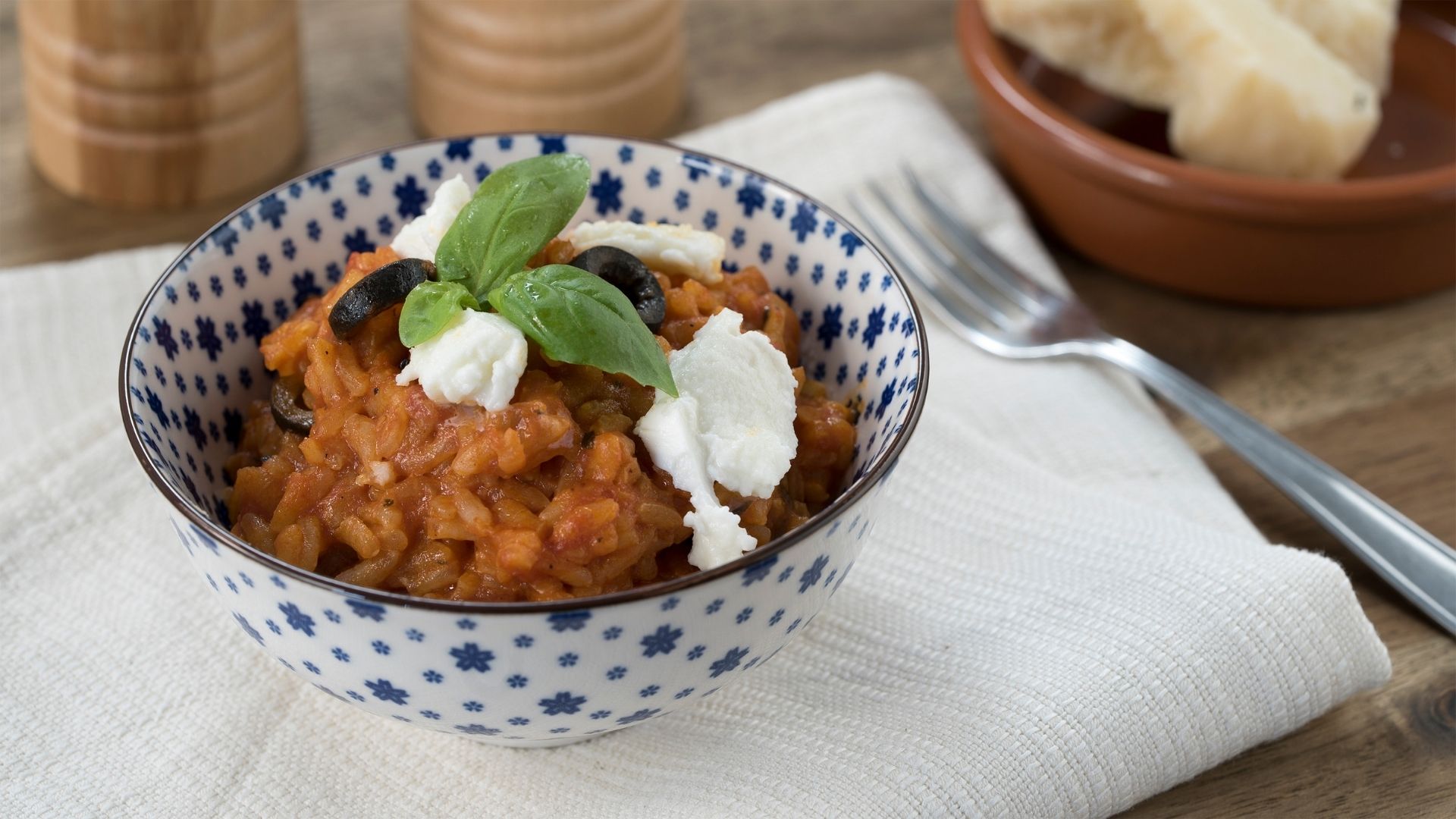 Alternativ-Text: Eine Schale gefüllt mit Tomaten-Risotto, garniert mit Oliven, Mozzarella und Basilikum